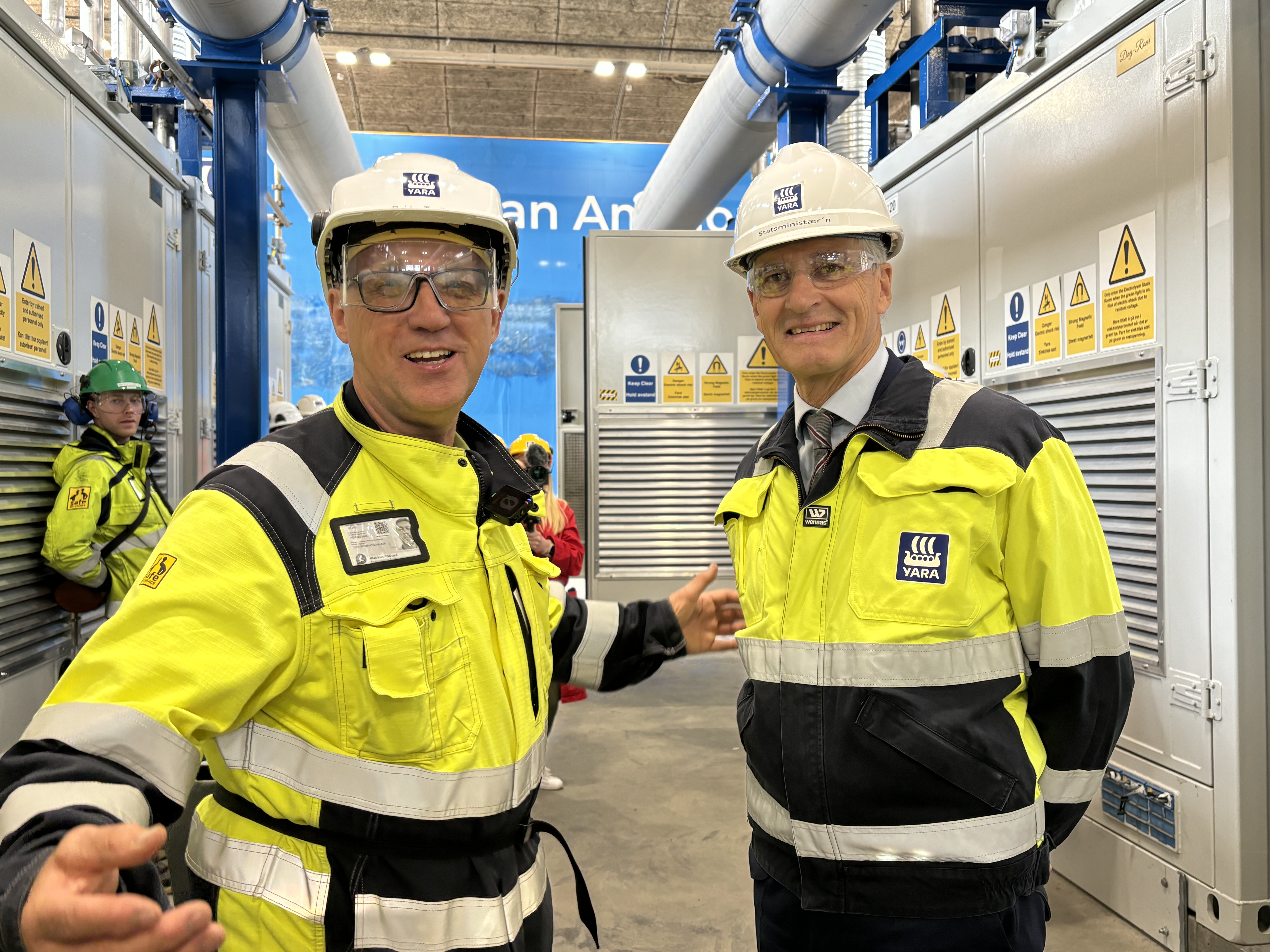 two men, wearing PPE, one is gesticulating while talking, posing, standing in a hydrogen plant. Opening ceremony.
