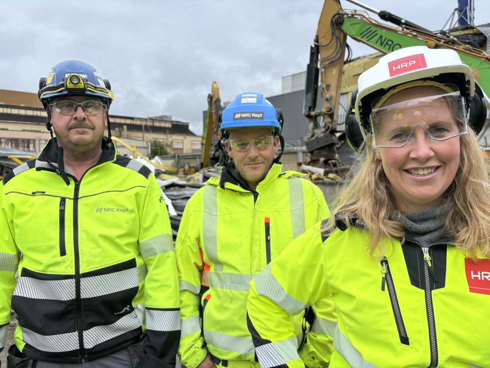 tre personer står ute på et anleggsområde, poserer, gule jakker og hjelmer og vernebriller. Anleggsmaskiner i bakgrunnen.