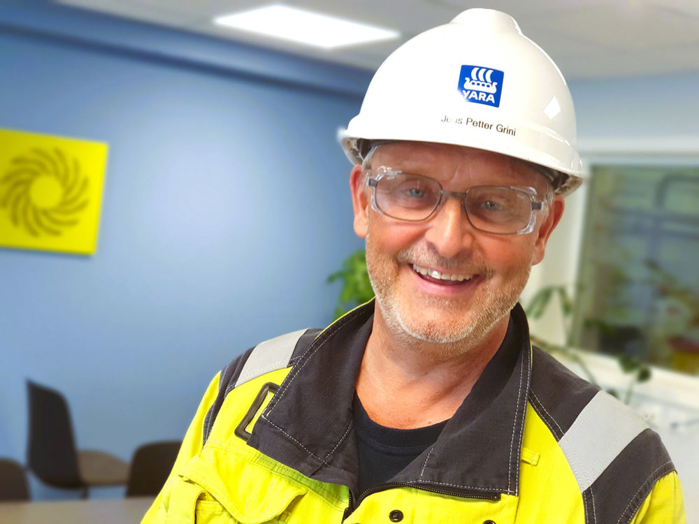 portrait of a smiling man wearing yellow jacket and white helmet, in a meeting room with blue wall