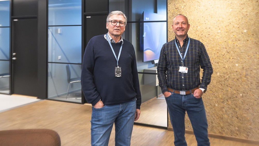 two men, posing, office premises, glass walls and black office doors in background.