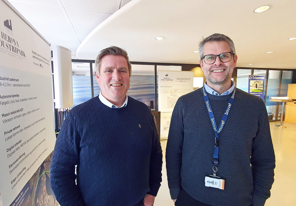 two men posing, wearing blue jumpers, standing in an open office space.