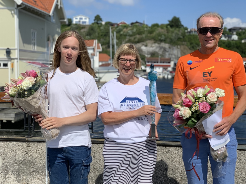 tre personer står på en lav bro, poserer, holder blomsterbuketter, blå himmel og sol, prisvinnere.