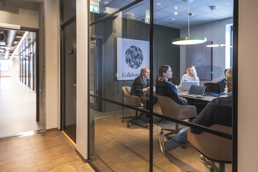 people sitting in a meeting room, glass walls, outside is a long corridor.