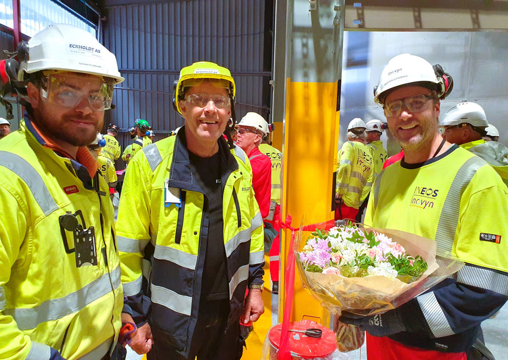tre menn i gult verneutstyr og hjelmer, en mann holder en blomsterbukett. Poserer, fabrikkhall.