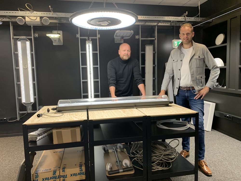 two men, posing, standing in a showroom environment, dark walls and a lot of lighting equipment.