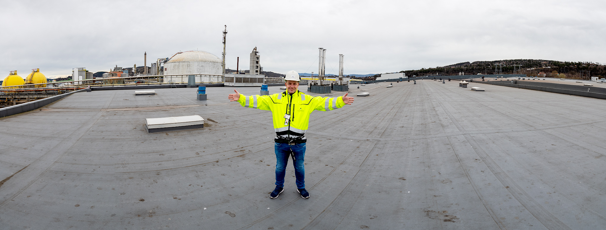 mann i gul jakke og hvit hjelm står med armene utstrakt på et stort tak, industrianlegg i bakgrunnen.