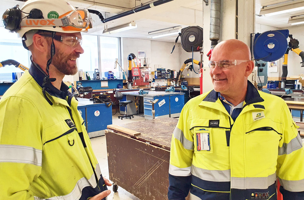 two men dressed in yellow work clothes talking in a workshop