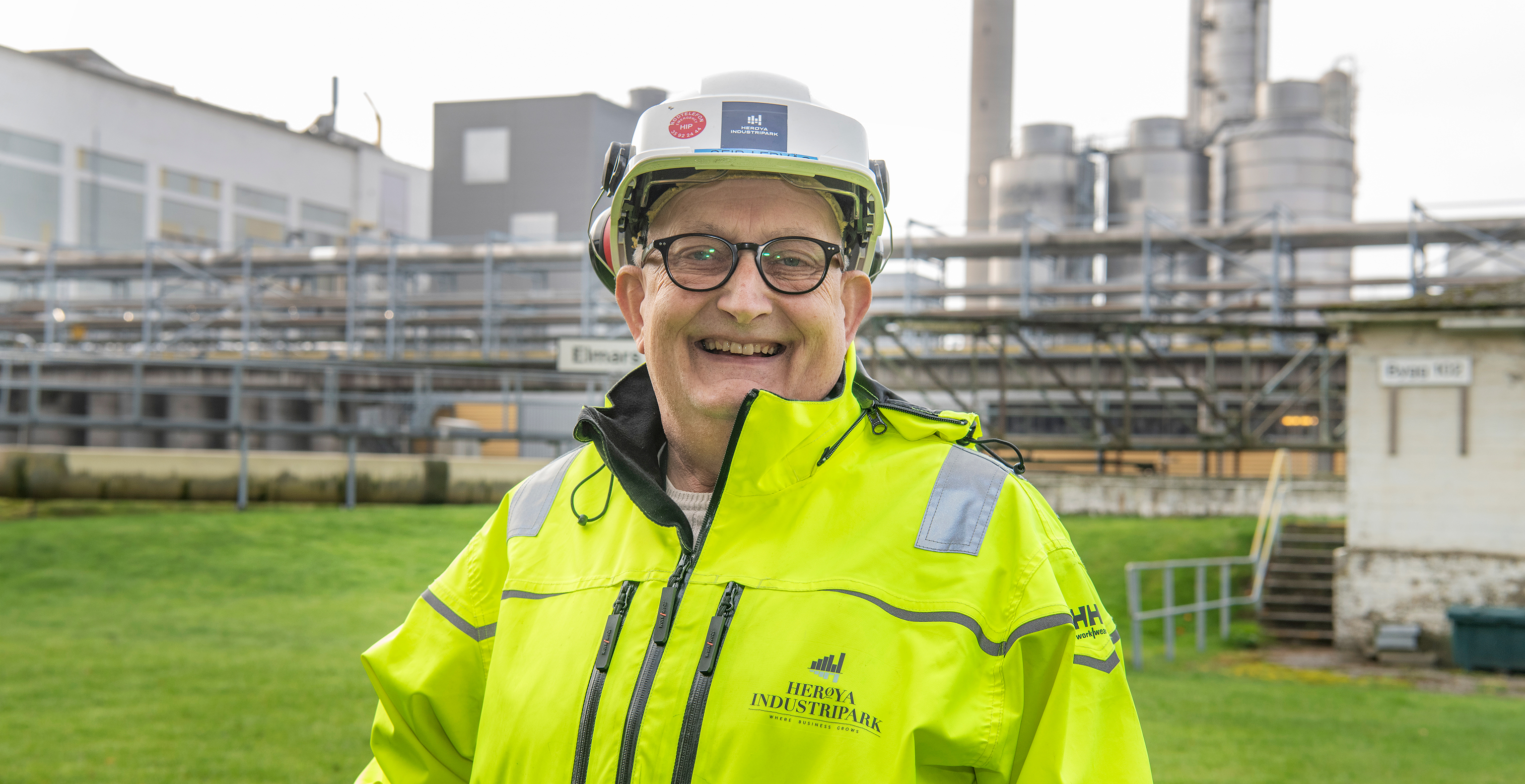 portrait of man, posing, smiling, yellow jacket, white helmet, green area, industrial site in background