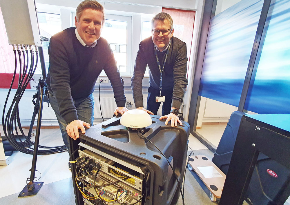 to menn poserer mens de lener seg mot en 5G-nettverksboks, i et rom med teknisk utstyr.