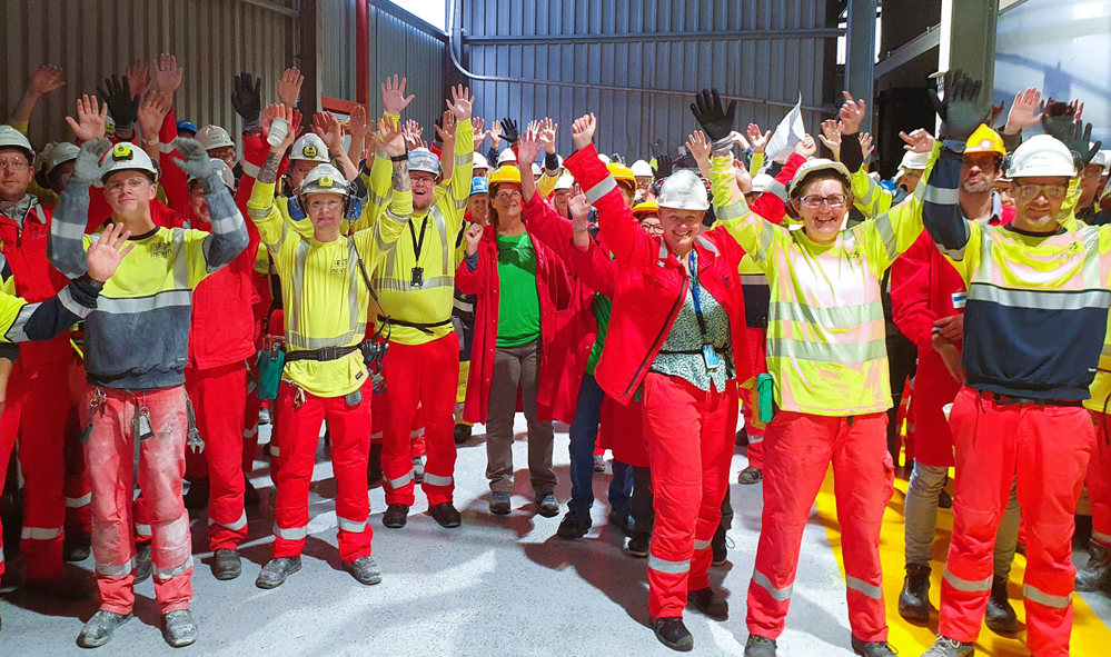 stor gruppe mennesker i rødt og gult arbeidstøy, armene i været, poserer, er glad
