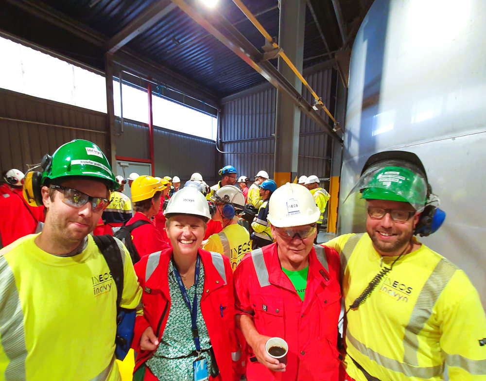 fire personer står tett sammen, smiler, poserer, ved siden an en stor ståltank i en produksjonshall