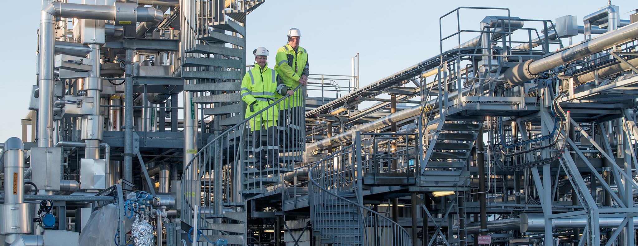 two men in yellow working clothes standing in a test rigg, a lot of pipes in steel