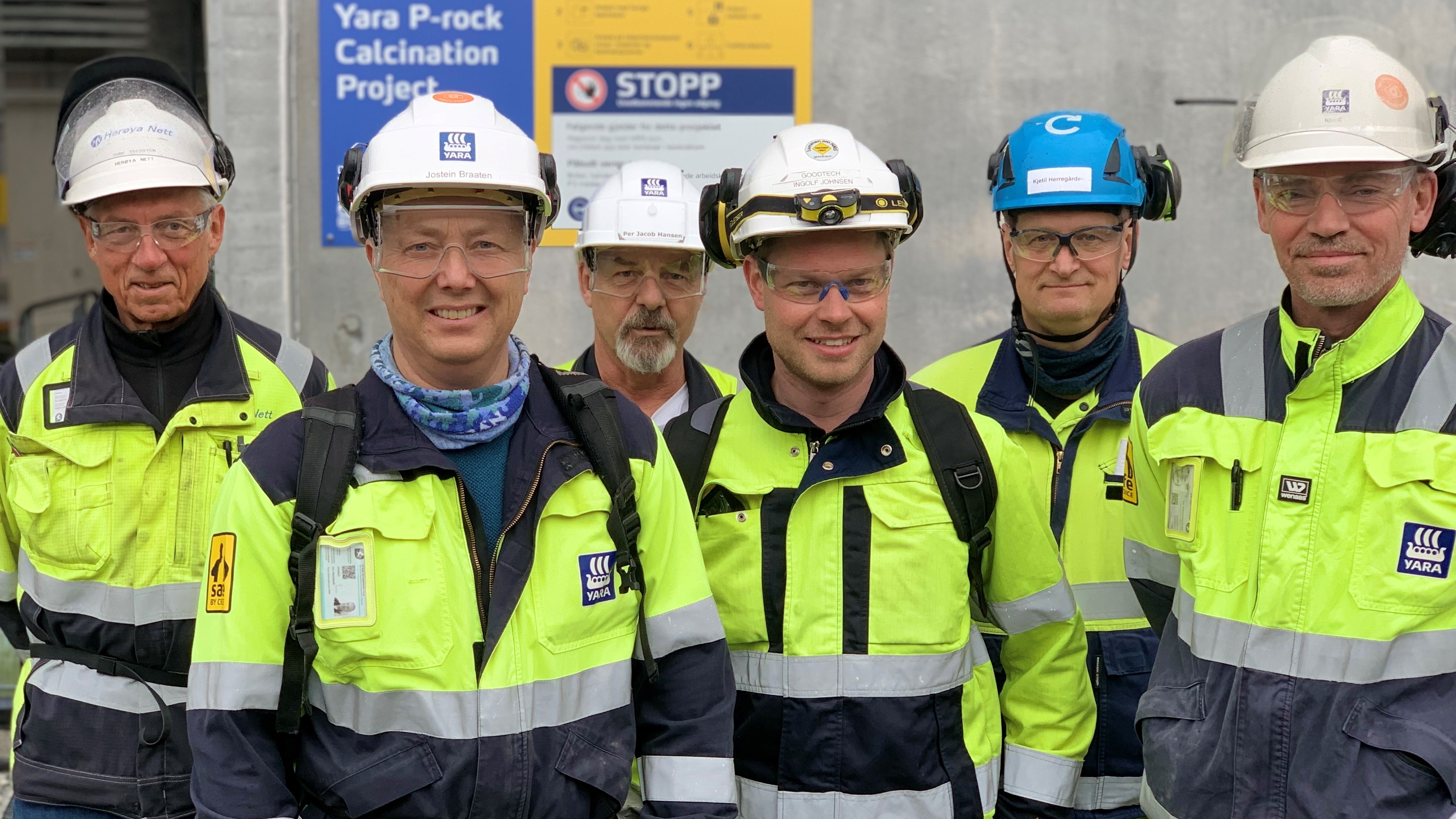 seks menn står sammen, gule og blå arbeidsklær, hjelmer, poserer inntil en betongvegg