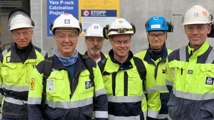 seks menn står sammen, gule og blå arbeidsklær, hjelmer, poserer inntil en betongvegg