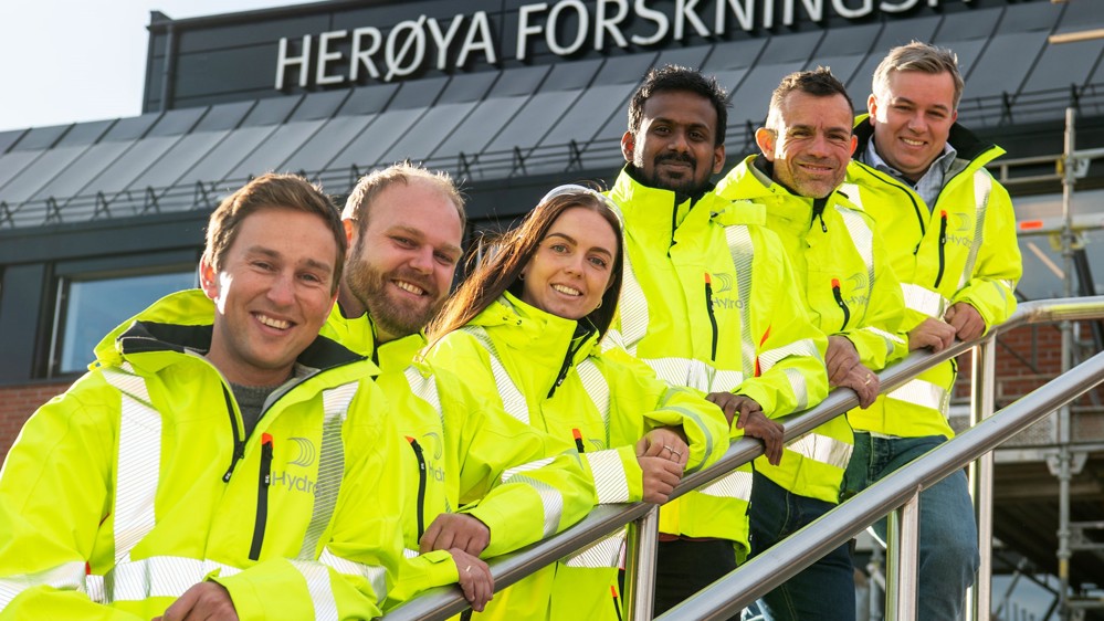 six people standing in stairs, leaning onto the rails, all wearing yellow jackets, posing, outside entrance to research park