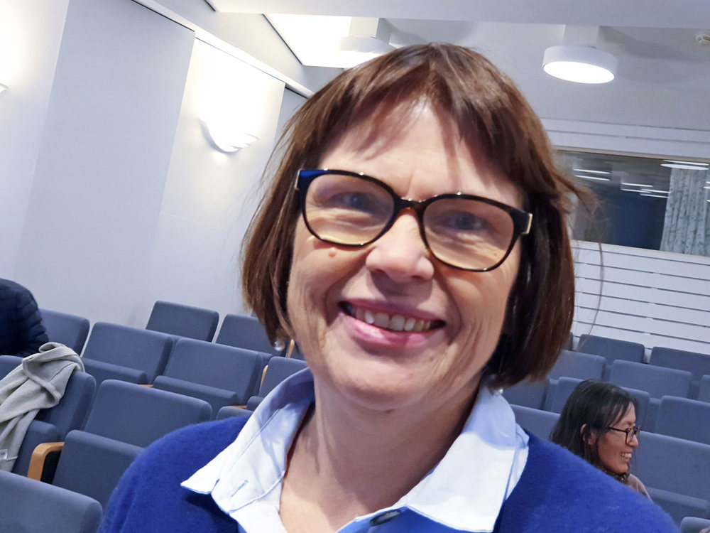 portrait of woman, smiling, posing, dark hair, dark rimmed glasses, auditorium, blue empty seats behind her.