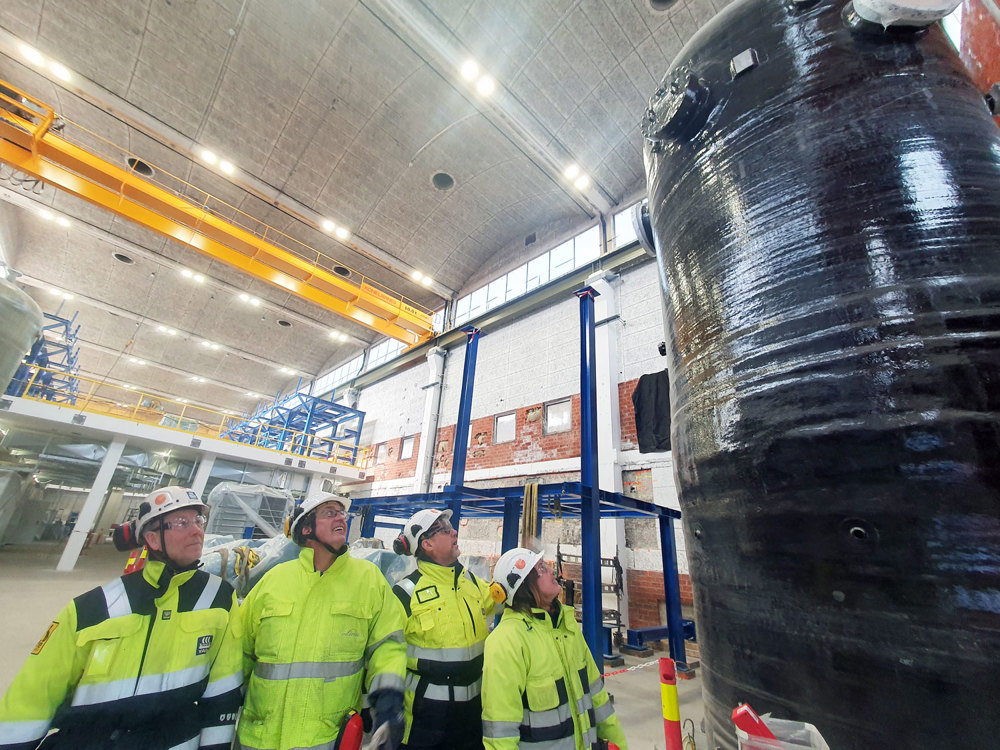 fire personer står på fabrikkgulv og ser opp på en stor oksygenseparator.