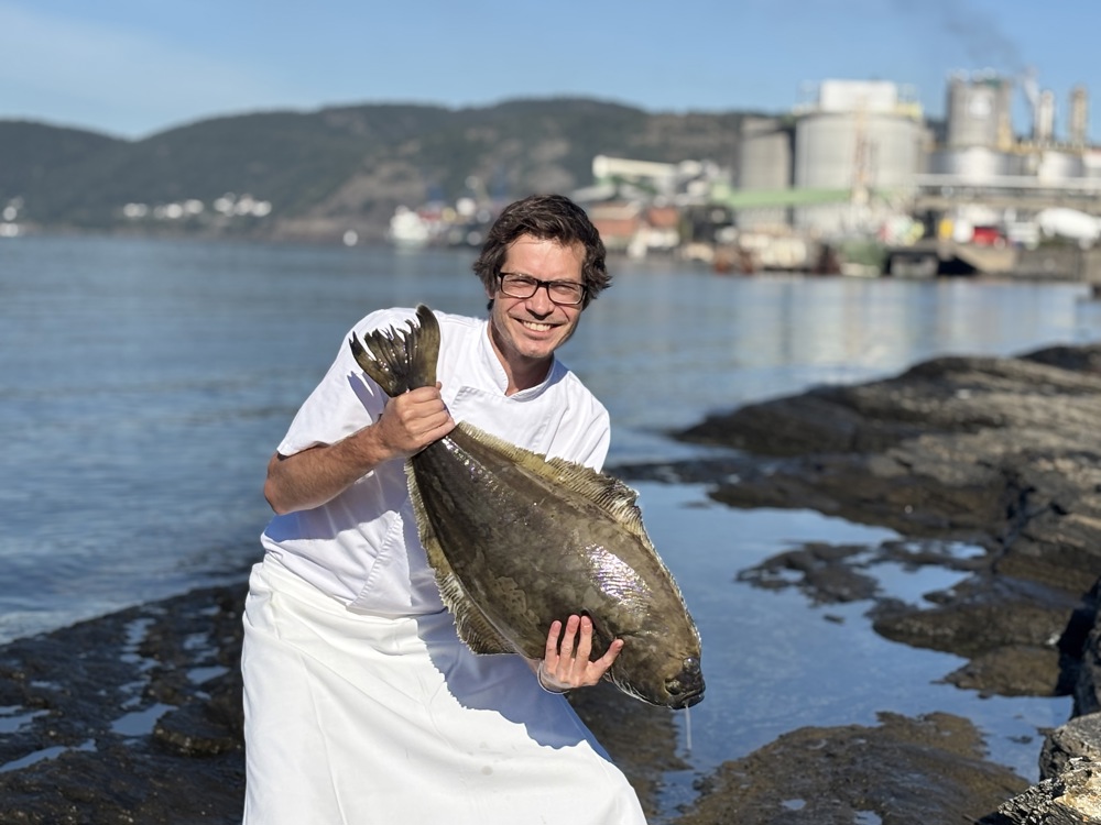 chef in white clothing, posing with a great fish in his hands, industry and sea in background.