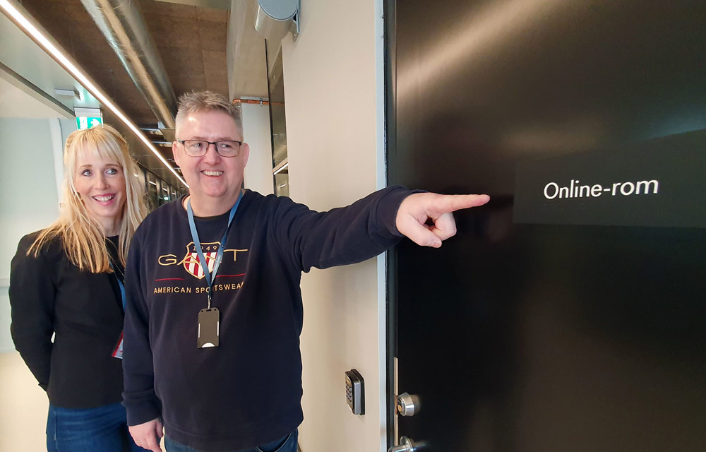 two persons standing outside a door in an office environment, one of the persons pointing at the door sign.