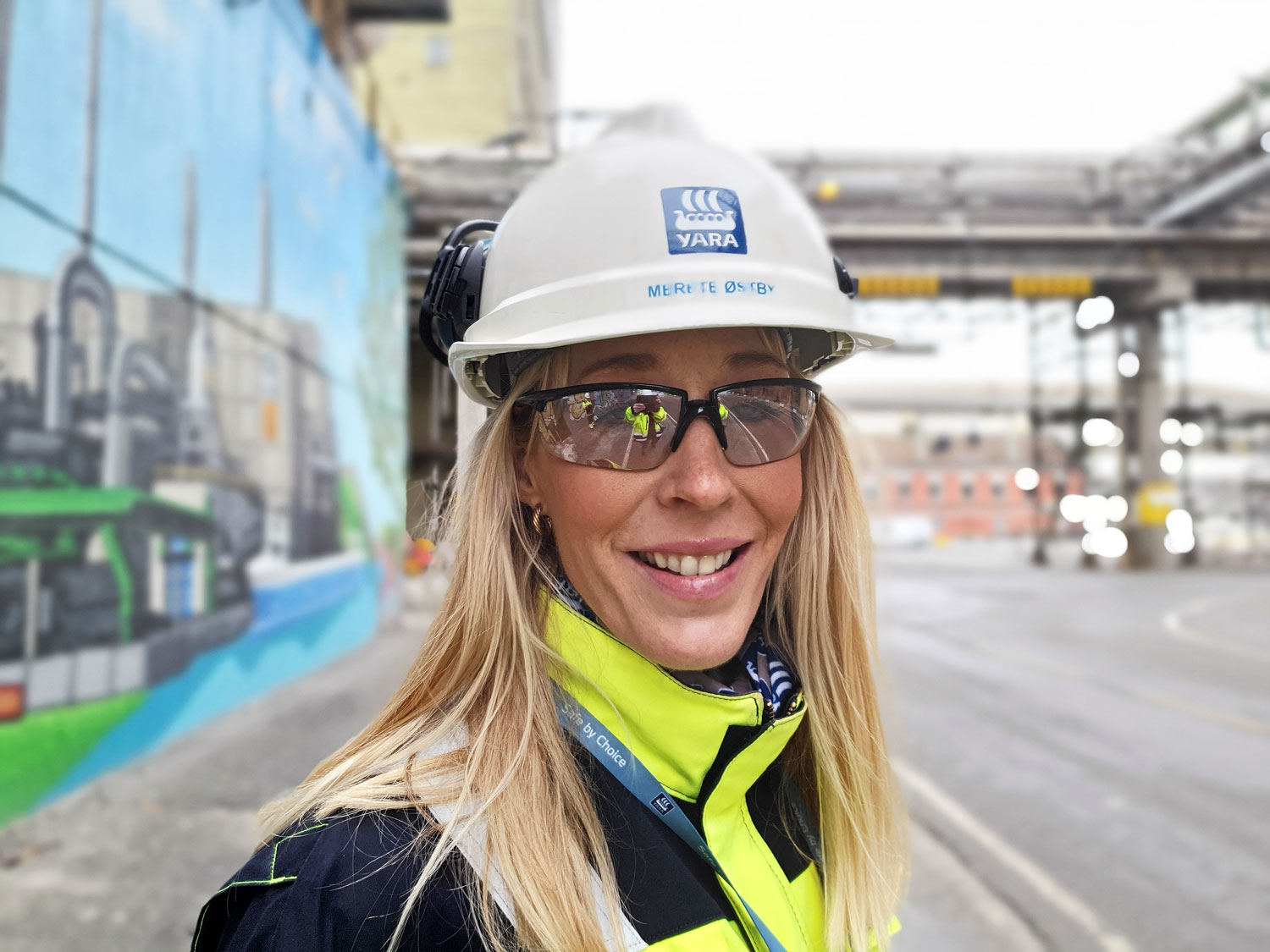 portrait of woman, posing, white helmet, yellow and blue jacket, long blond hair, goggles. She is standing in a street in an industrial park. Decorated concrete wall behind her.