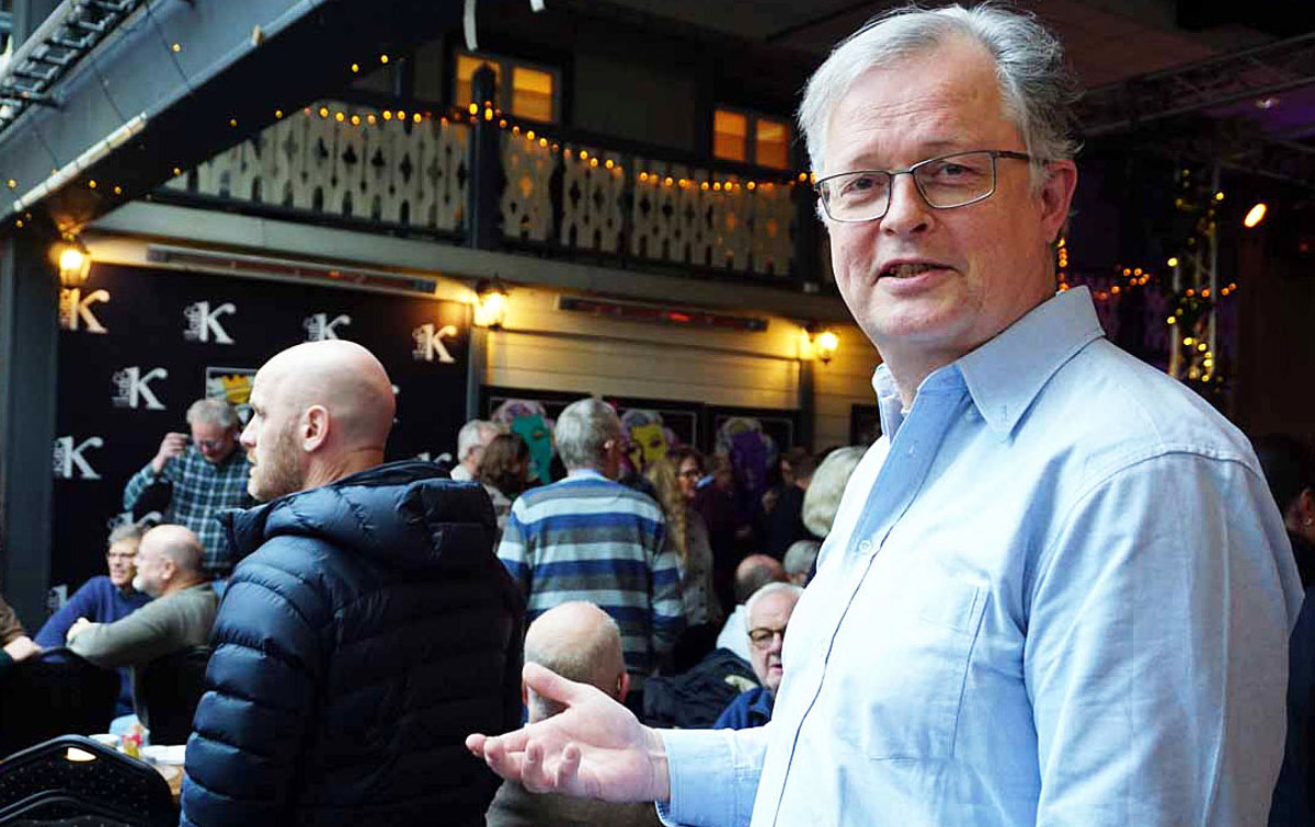 portrett av mann, står i lokale med mange mennesker, som sitter og står