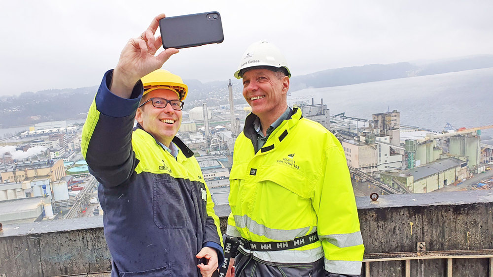 to menn kledd i profileringstøy og hjelmer, står på toppen av et høyt bygg, den ene tar en selfie av dem begge