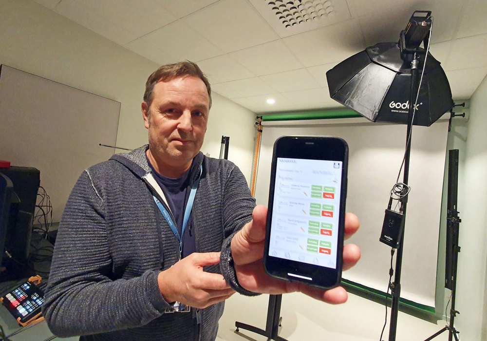 man, posing, and displaying his mobil and showing an app. Studio with lamps and monitors in background.