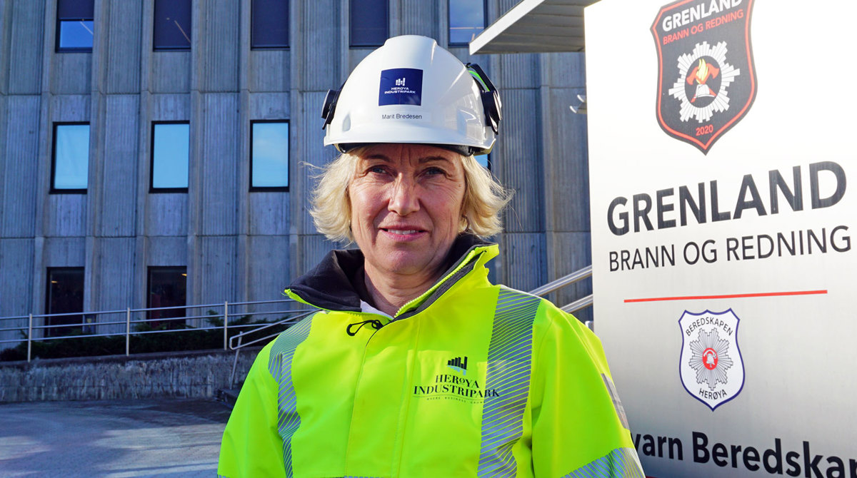 portrett, kvinne, poserer, gul jakke, hvit hjelm, firmaskilt bak, og stort kontorbygg i bakgrunnen