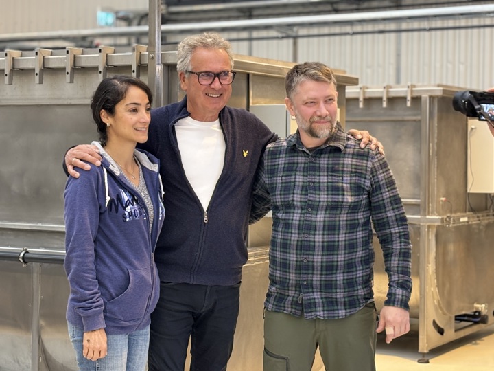 tre personer står sammen armene på skuldrene til hverandre, poserer, smiler, stor lagerhall, produksjonsutstyr