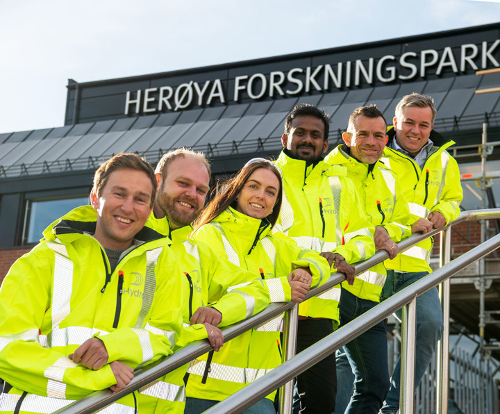 Six people, dressed in yellow jackets, pose, stand up a flight of stairs and lean against the railing outside a brick building. On the top of the building, large letters that read Herøya Forskningspark are partially displayed