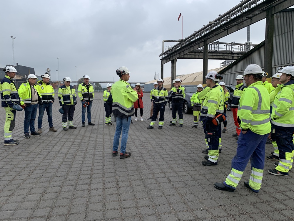 gruppe mennesker, alle kledd i gule og blå arbeidsklær, hører når en person som står i midten snakker