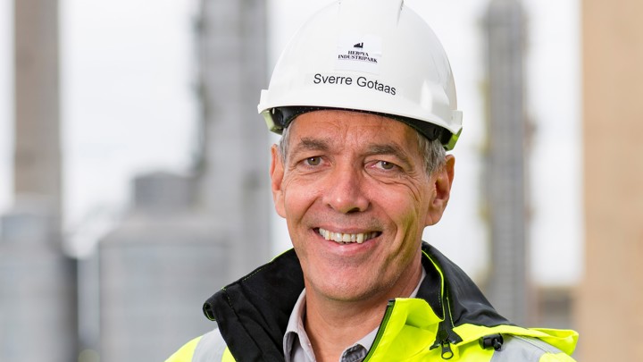 Portrait of a man wearing yellow jacket and a white helmet, industrial park below in background.