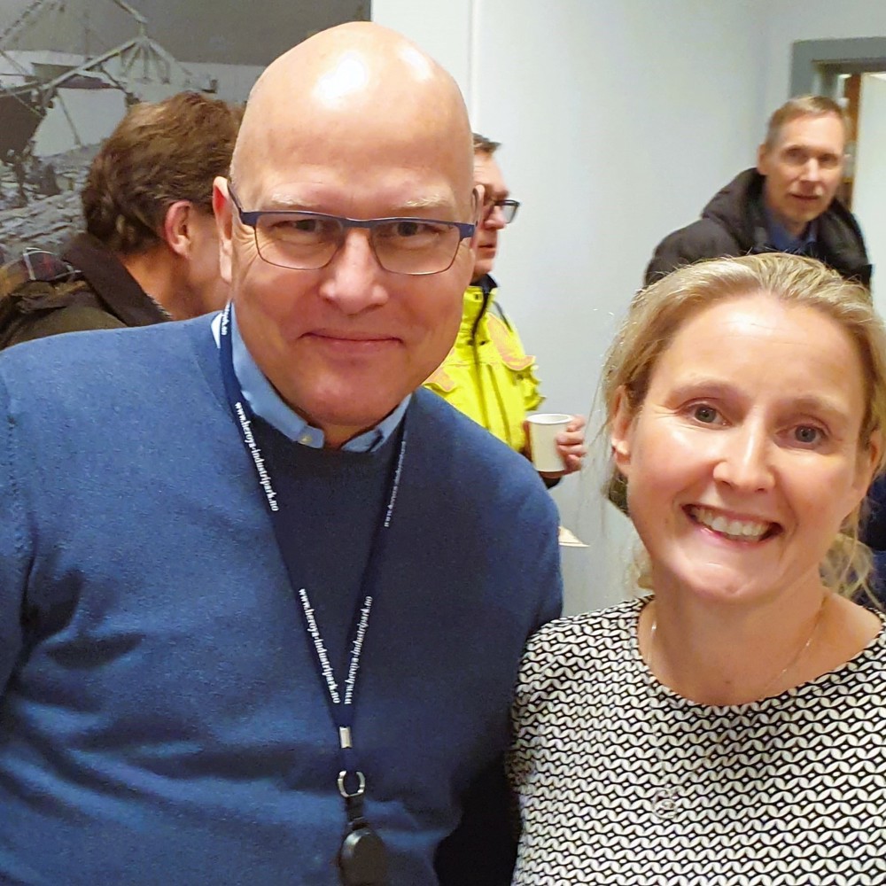 man and woman standing close next to eachother, people behind them, meeting room