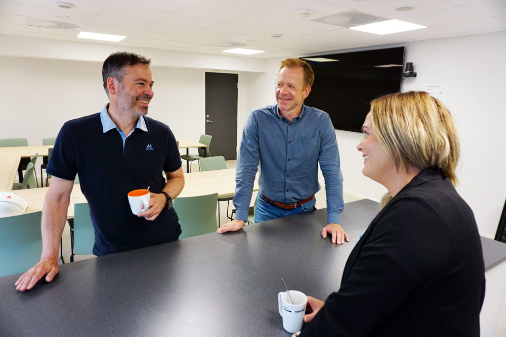 tre personer med en kaffekopp hver står ved et bord, samtale, kantine