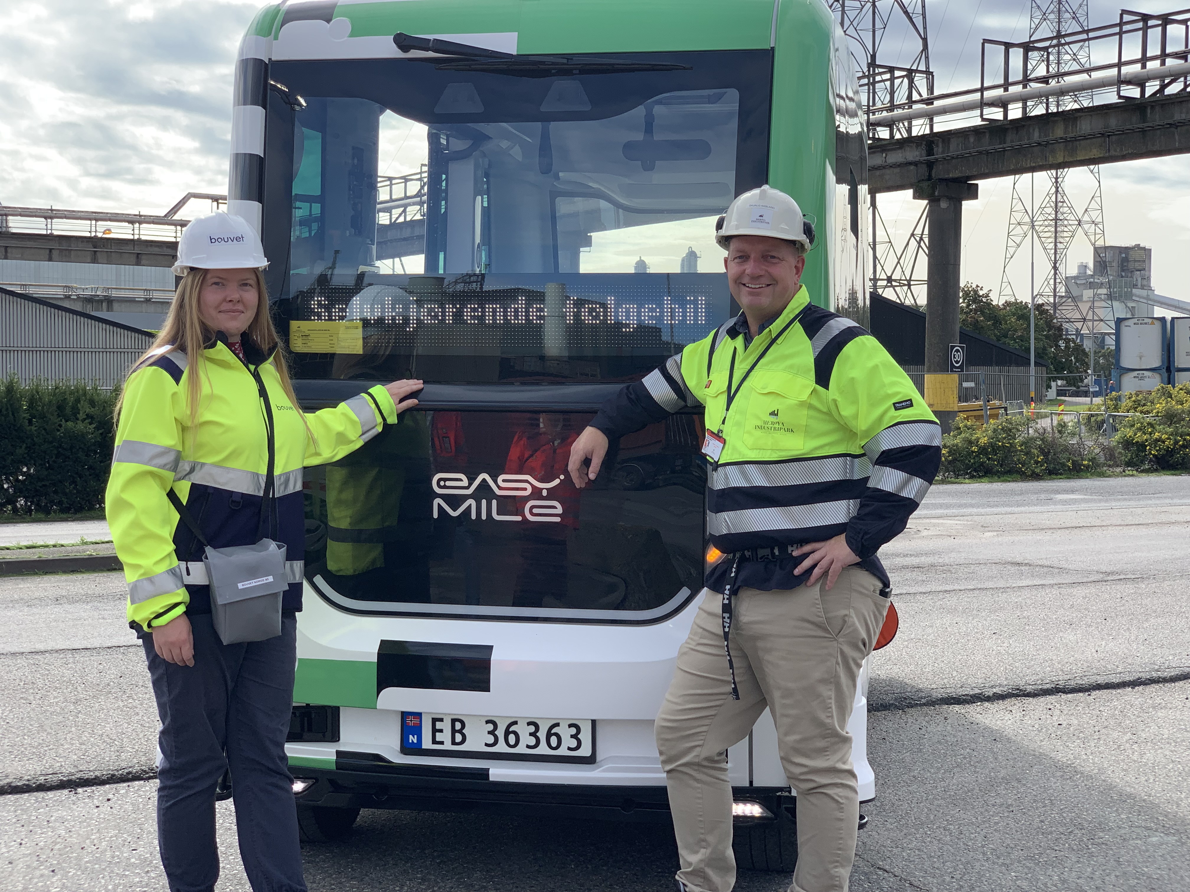 two persons posing in front of an autonomous white mini bus, located outside and in an industrial environment.