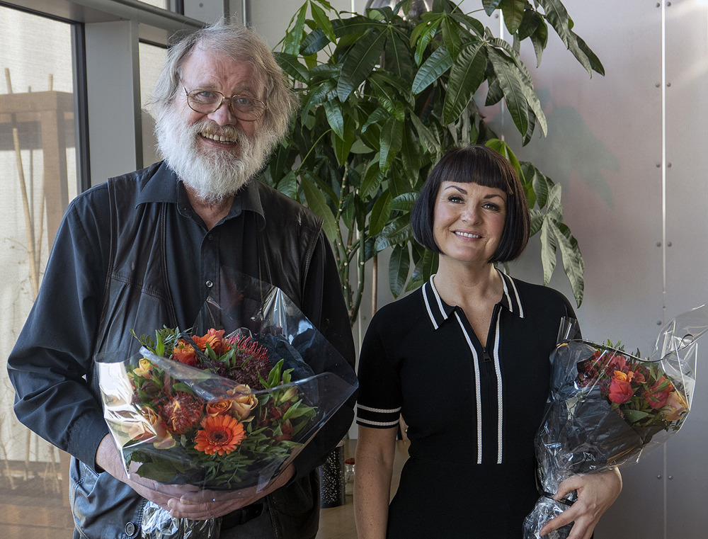 mann og kvinne står sammen, poserer, med hver sin blomsterbukett. Mann har grått hår og skjegg og kvinnen har mørkt halvlangt hår. De står i et hjørne i et rom med vinduer og grønn plante bak.