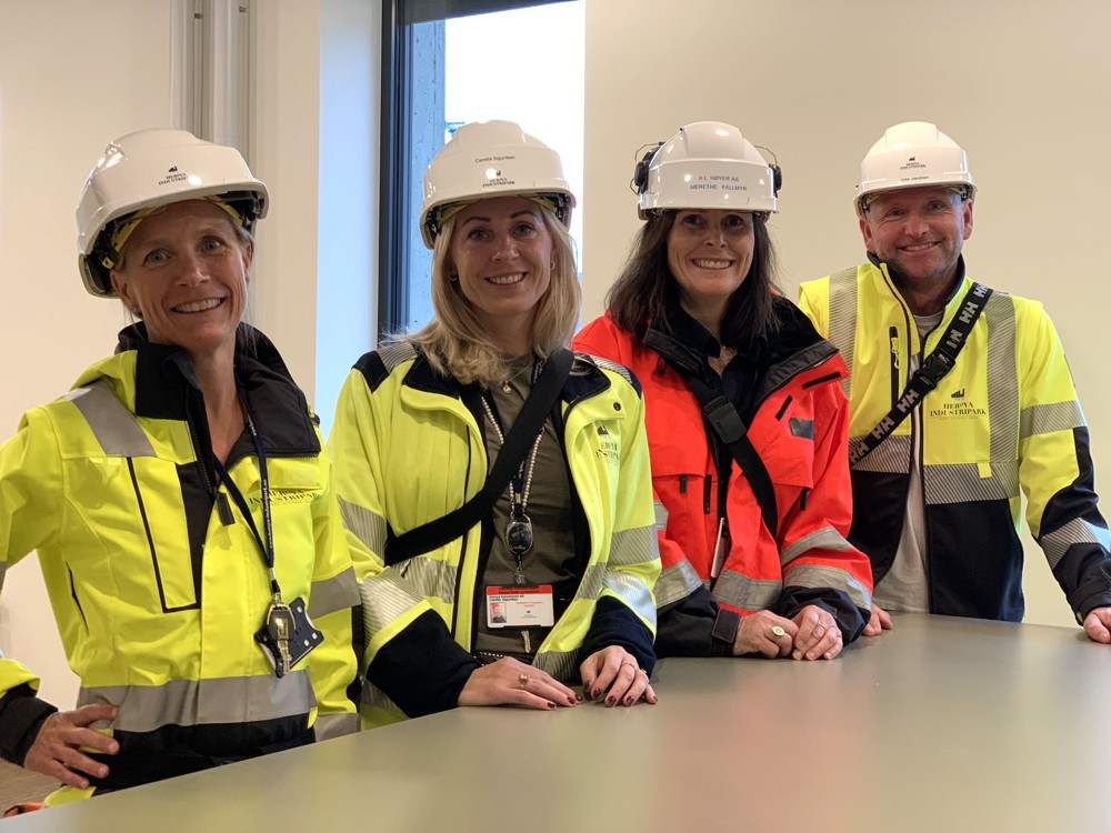 Four people standing at a bench, all in yellow and orange work jackets, white helmets, office environment.