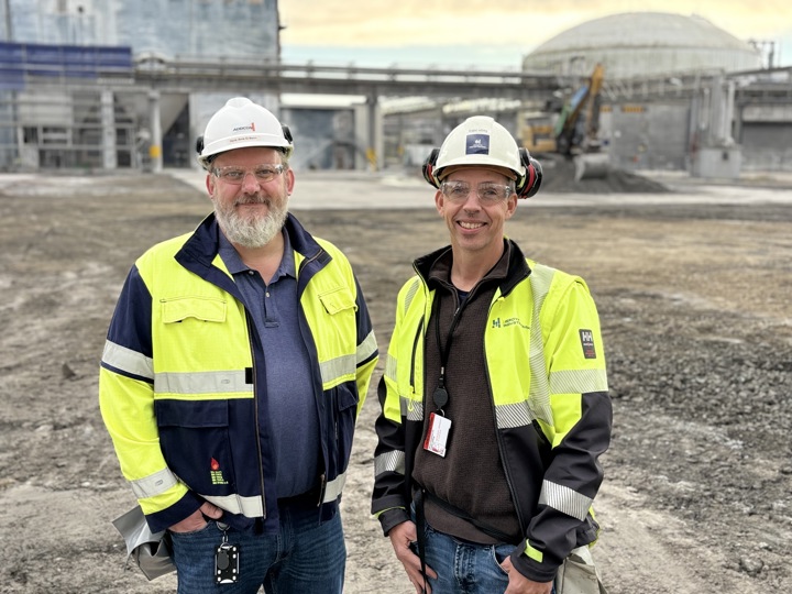 to menn, poserer, arbeidsklær, hvite hjelmer, industritomt i bakgrunnen