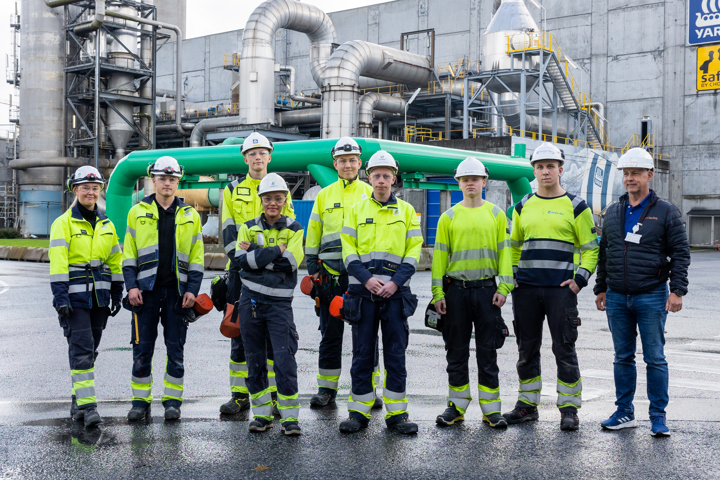lærlinger står på rad, i gule jakker og blå bukser, poserer, fabrikkanlegg i bakgrunnen