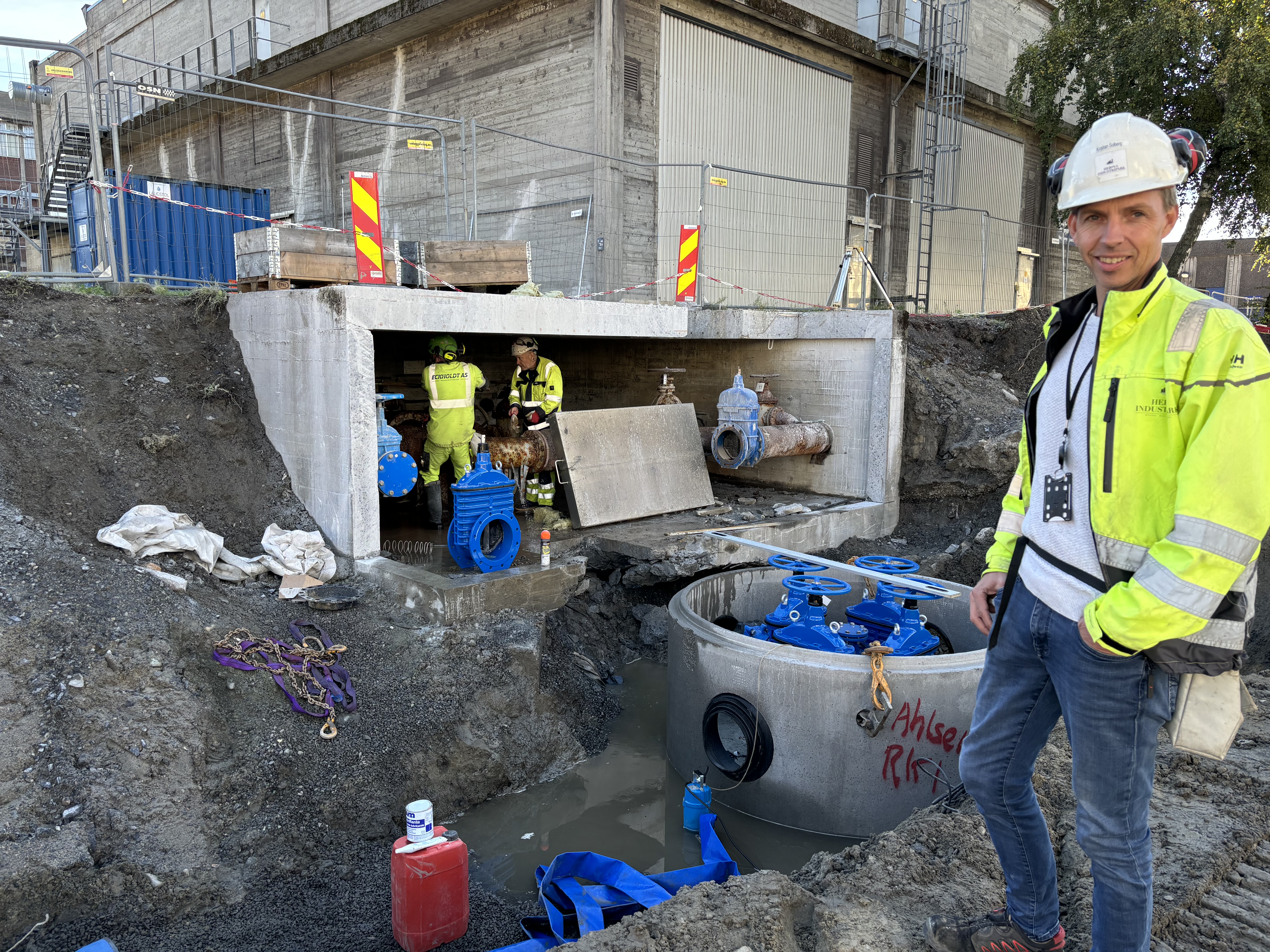 mann i gul jakke og hjelm står foran grøft med ny vannpumpe i kum