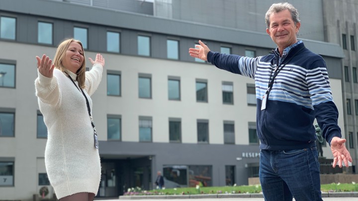 kvinne og mann poserer utenfor kontorbygg med armene ut, pekende mot bygget