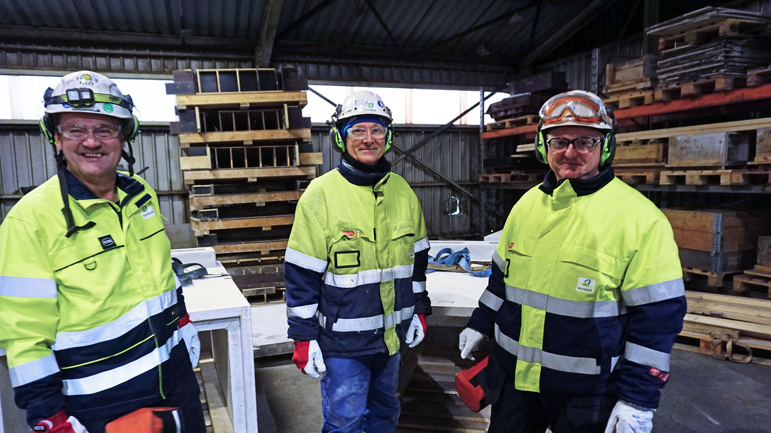 three persons, posing, wearing yellow jackets, white helmets, goggles and hearing protection, standing in a workshop