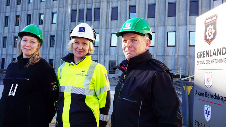 three persons, posing, wearing PPE, concrete builing with lots of windows in background, pylon with business logoes next to the people.