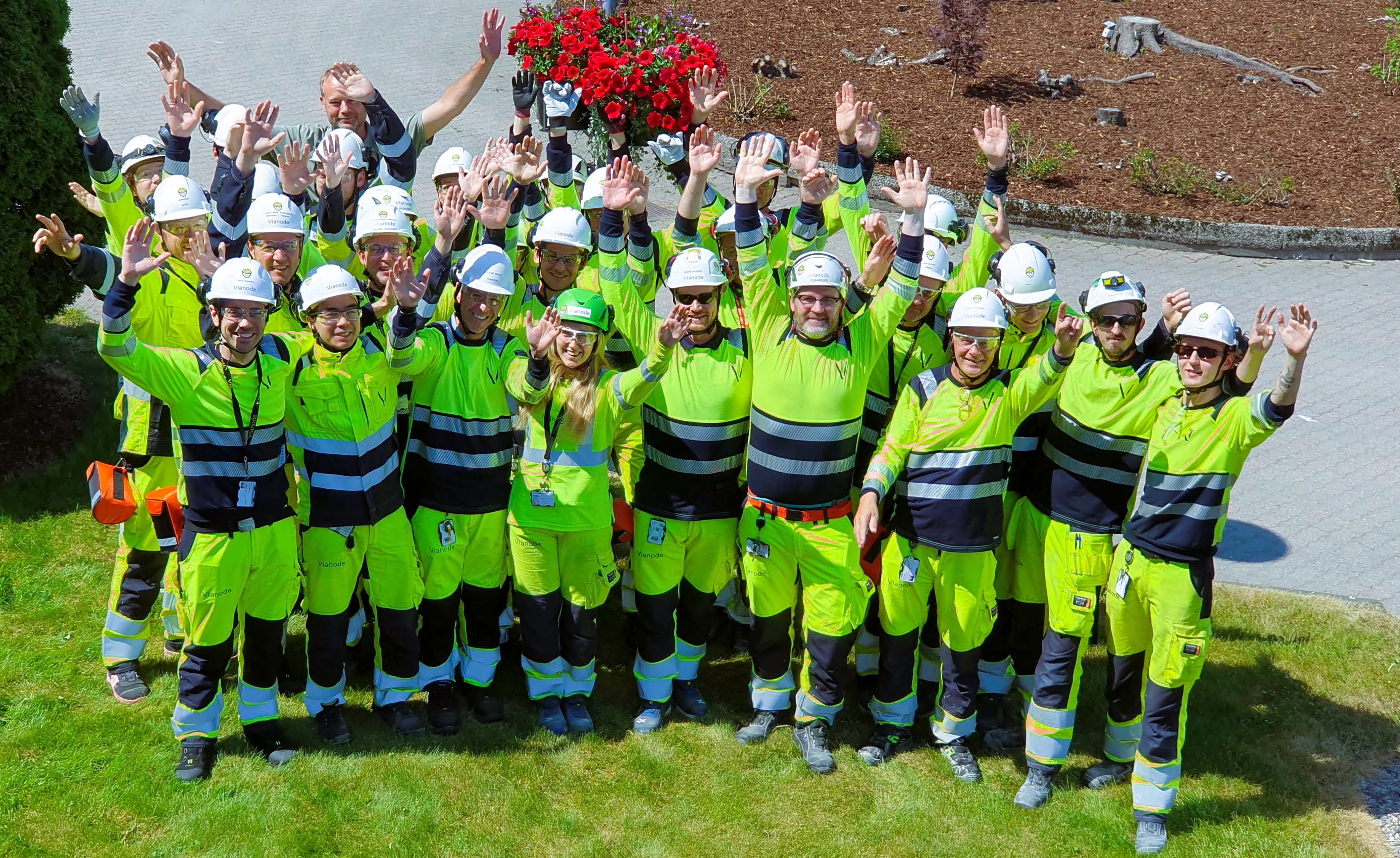 group of new employees, about 30 people, stand close together and look towards the photographer who is at the height, they stretch their arms in the air. All dressed in yellow and blue work clothes and helmets with chin straps.