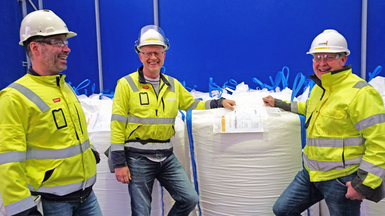 three men posing, wearing PPE, standing close to big bags of products, all smiling.