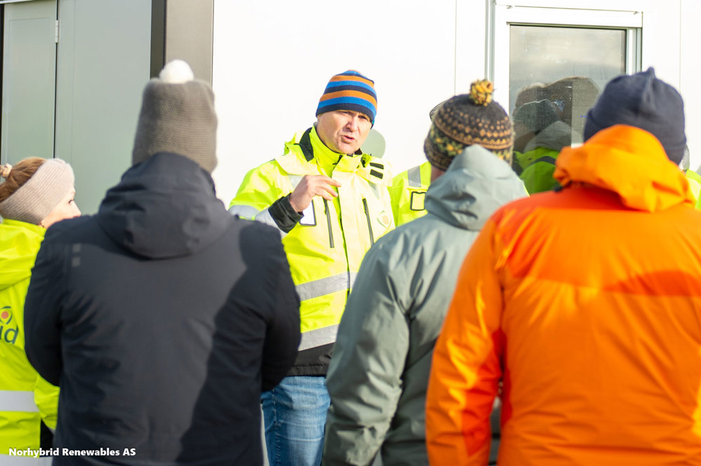 mann i gul jakke står og snakker til en gruppe mennesker, alle kledd i tjukke jakker og lue, står med rygge til kamera. Han som snakker står inntil en husvegg, ved vindu.