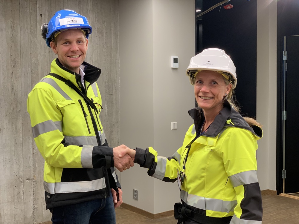 A man and a woman, posing, shaking hands, thanking for the delivery, wearing yellow jackets and blue and white helmets, office environment.