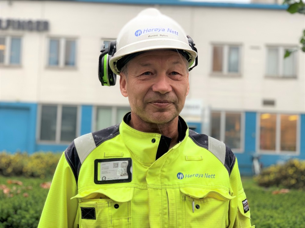 portrait of a man, wearing personal protective equipment, behind him is a blue and white building and a green hedge.