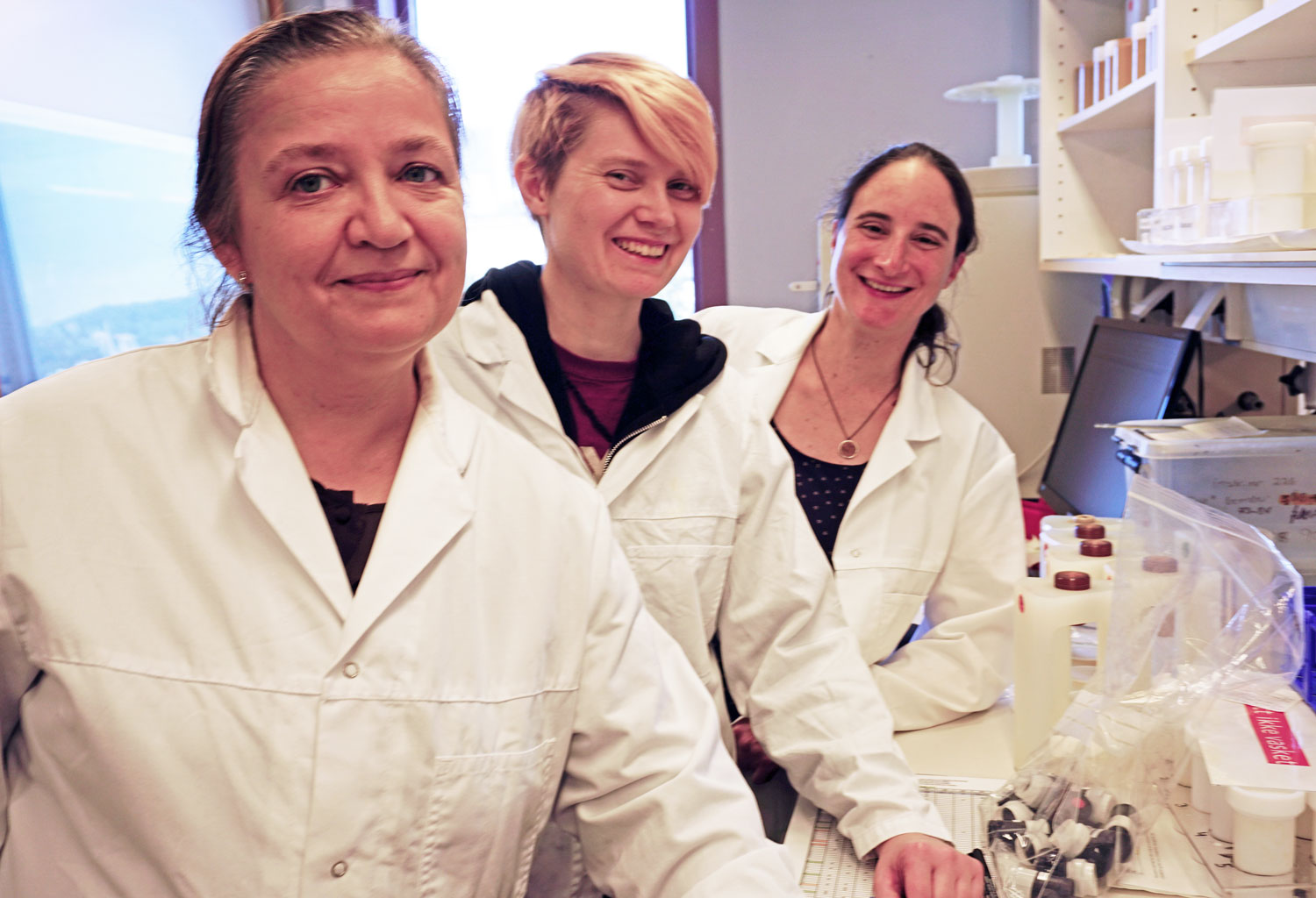 Three women, white lab coates, goggles, posing, leaning on to a work table in a laboratory