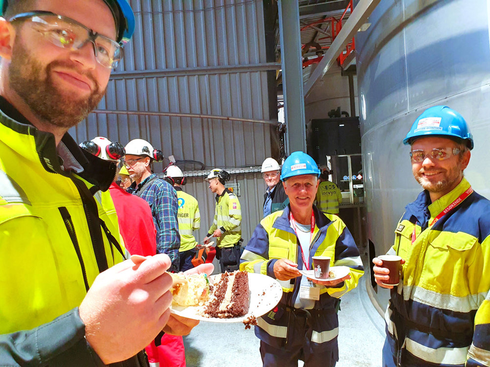 tre menn, gule jakker, blå hjelmer, poserer og spiser kake og har kaffekopp. Fabrikkhall.